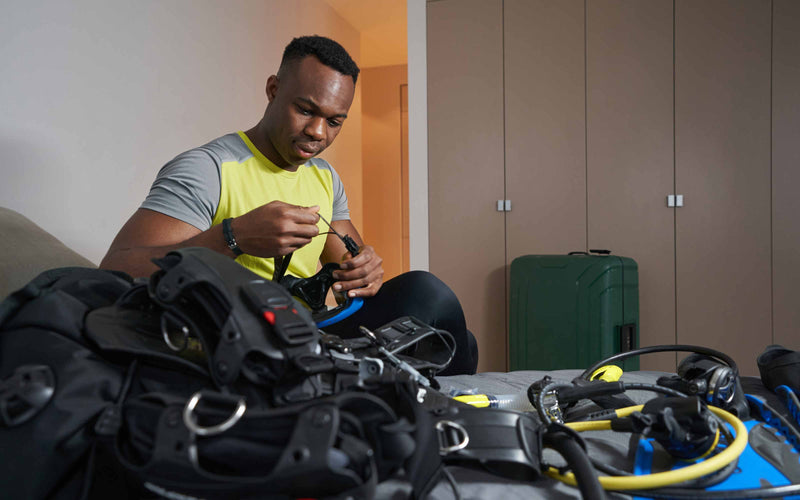Person sitting on a bed assembling scuba diving gear, with various equipment spread out and a green suitcase in the background
