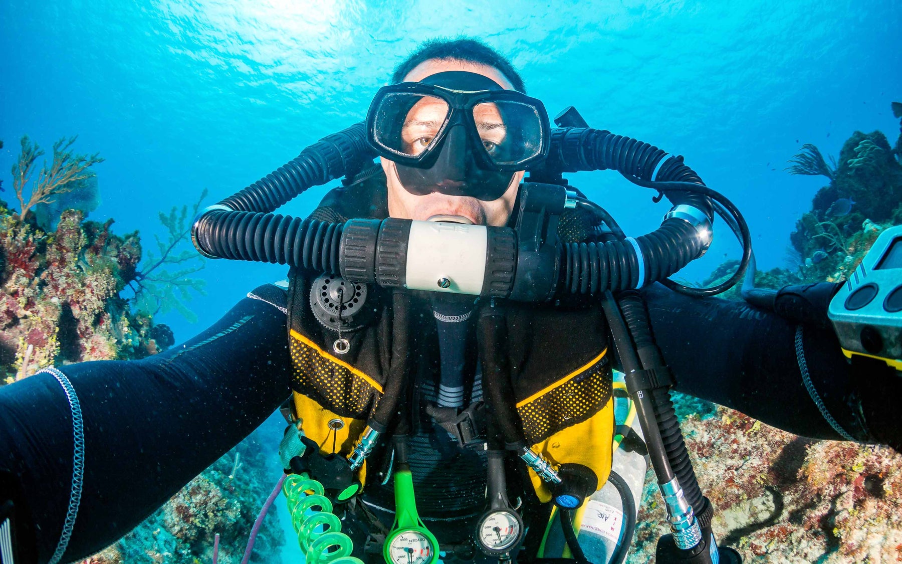 Underwater close-up of a scuba diver wearing a full-face mask and rebreather system, surrounded by coral and marine life
