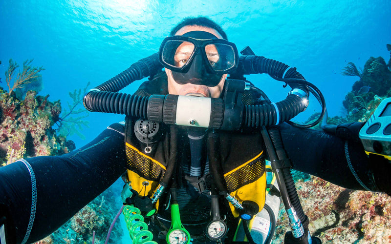 Underwater close-up of a scuba diver wearing a full-face mask and rebreather system, surrounded by coral and marine life